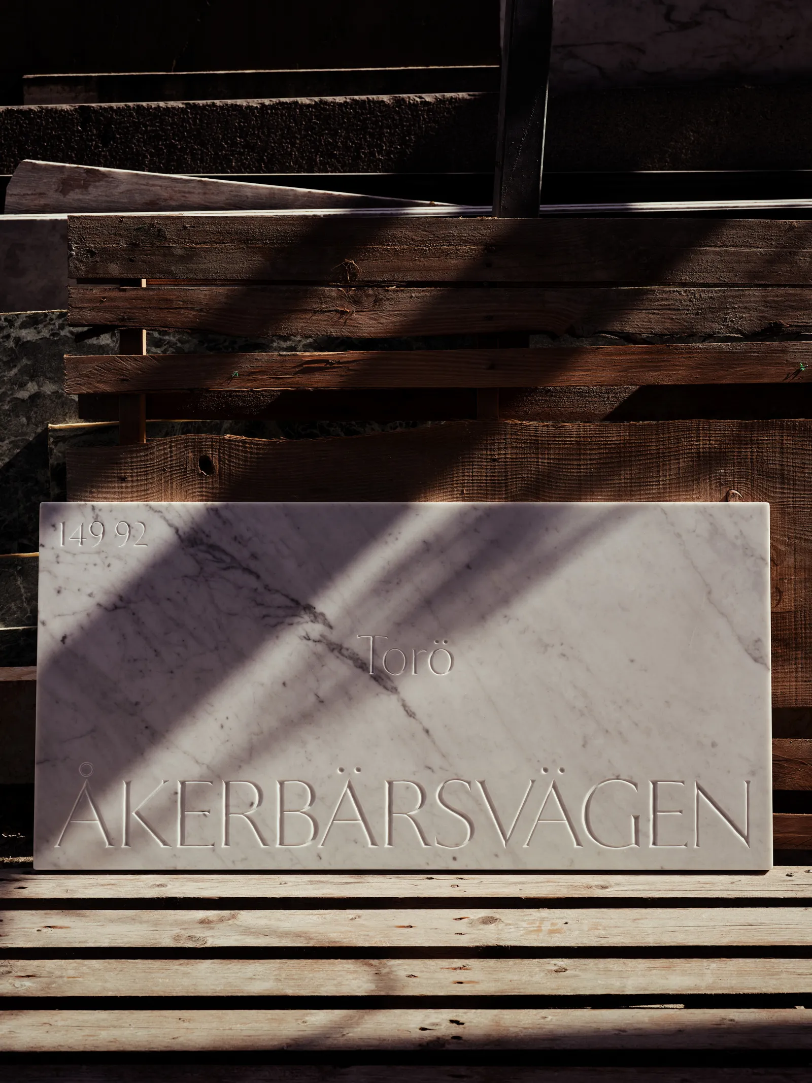 The Åkerbärsvägen marble sign leaning against wooden pallets in raking outdoor sunlight.