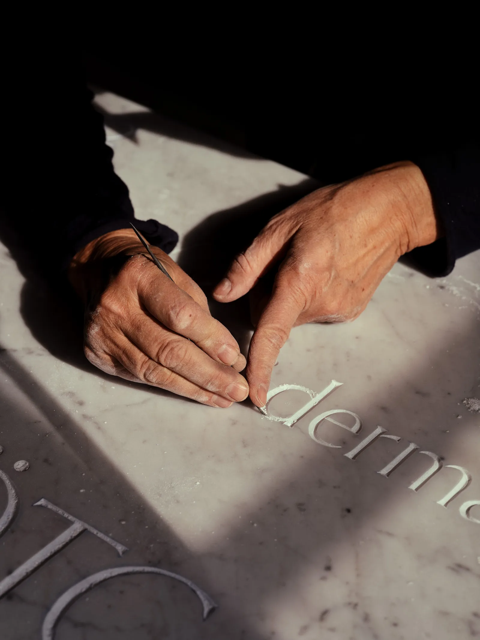 A carver’s hands guiding a chisel through a lowercase letter, mid-cut into a marble slab.